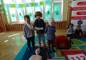 Chłopiec z temblakiem , dziewczynka z bandażem na ręku i chłopiec z bandażem na głowie podczas szkolenia z pierwszej pomocy przedmedycznej